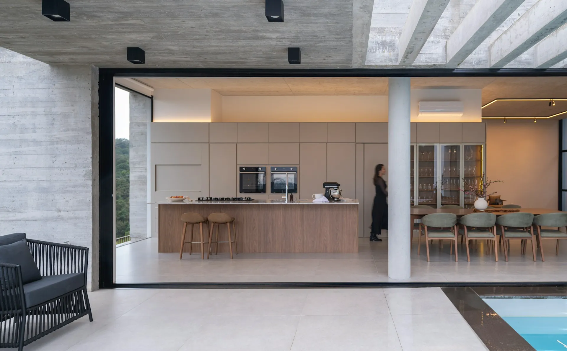 Modern open-concept kitchen and dining area with a large island, minimalist cabinetry, floor-to-ceiling openings, and a poolside patio.